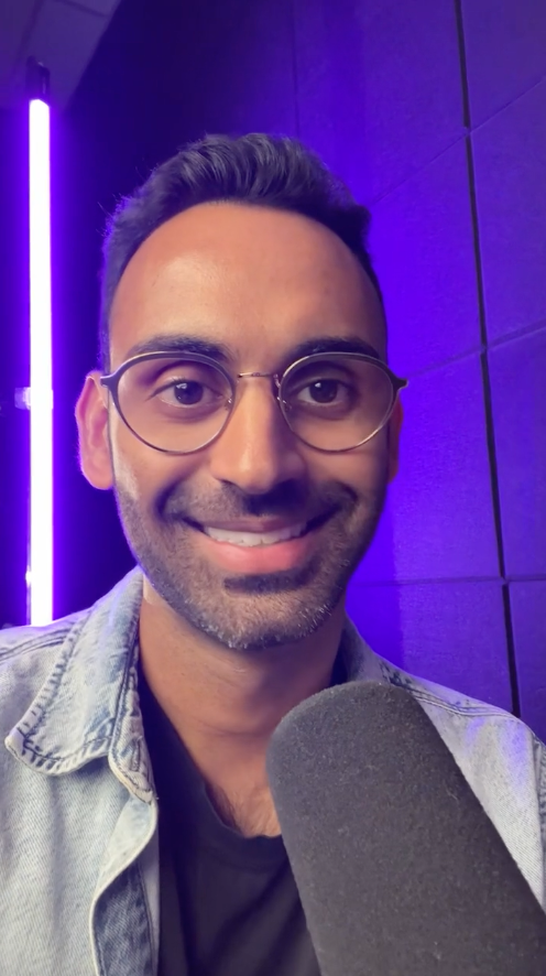 The Content Factory A person wearing glasses and a denim jacket smiles at the camera, holding a microphone in what looks like The Content Factory's podcast studio, with a purple neon light in the background. In Austin, Texas