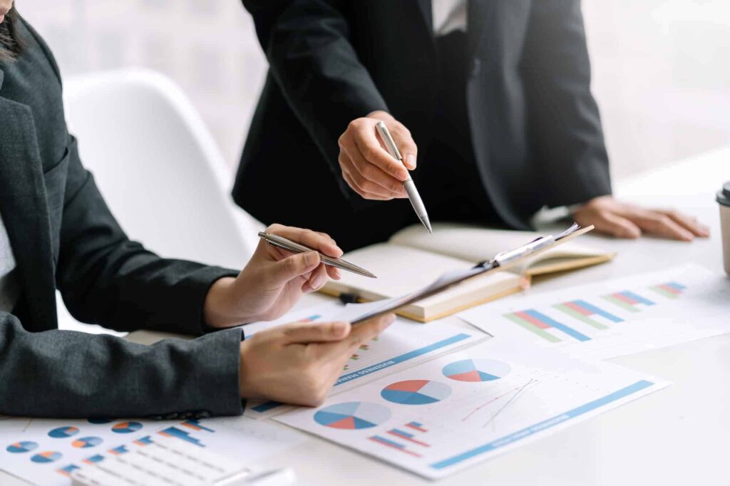 The Content Factory Two people in business attire review and discuss charts and graphs on documents laid out on a table in The Content Factory, using pens to point to specific data points. In Austin, Texas