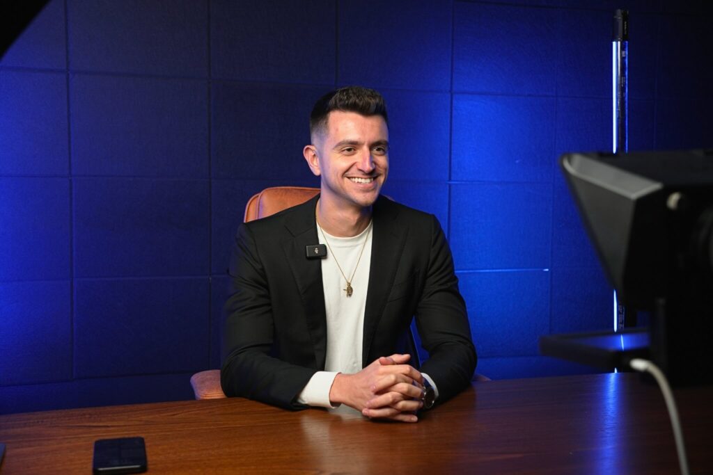 The Content Factory Person sitting at a desk in a recording studio, wearing a black blazer and white shirt, smiling at a camera. Background features a blue wall and studio lights. In Austin, Texas