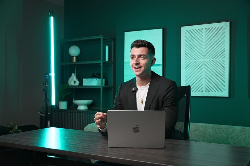 The Content Factory Person in a black blazer sitting at a desk with a laptop, speaking in a well-lit room featuring teal walls and modern decor. In Austin, Texas