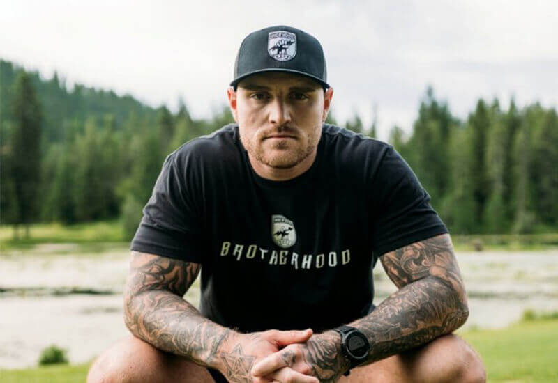 The Content Factory A man with tattoos sits outdoors on grass, wearing a black "BROTHERHOOD" t-shirt and a black cap with a logo, with trees and water in the background. In Austin, Texas