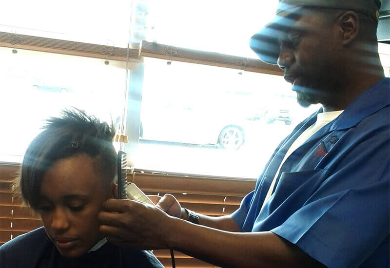 The Content Factory A barber in a blue jacket gives a haircut to a seated person using electric clippers near a window with sunlight streaming in. In Austin, Texas