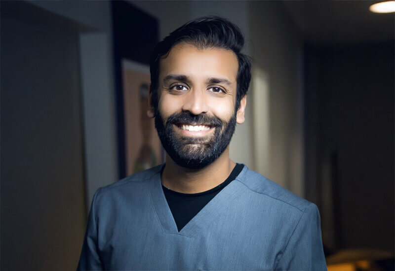 The Content Factory A man with a beard and dark hair, wearing gray medical scrubs, smiles while standing indoors in a softly lit hallway. In Austin, Texas