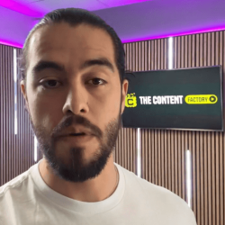 The Content Factory A man with a beard and tied-back hair wearing a white shirt is speaking in front of a wall with vertical wooden slats and a screen displaying "THE CONTENT FACTORY" logo, emphasizing the vibrant content creation scene in Austin, Texas (ATX). In Austin, Texas