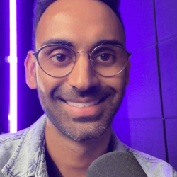 The Content Factory A person wearing glasses and a denim jacket smiles at the camera, holding a microphone in what looks like The Content Factory's podcast studio, with a purple neon light in the background. In Austin, Texas
